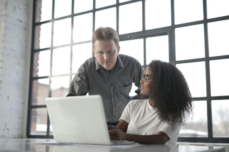 Professional adult male entrepreneur browsing netbook with young female African American colleague and discussing startup project during office work in workplace near big window